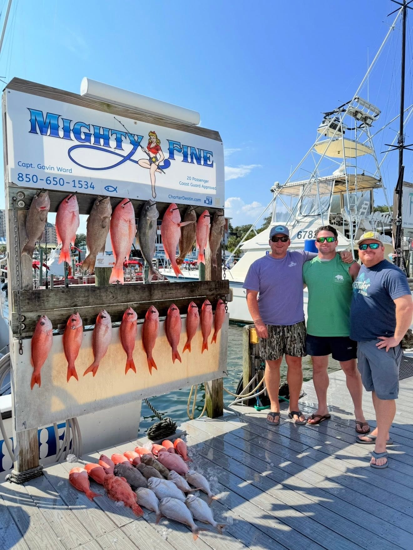 Three smiling anglers at the Destin dock beside the Mighty Fine catch board, hung with red snapper, grouper, amberjack, and a row of vermilion snapper